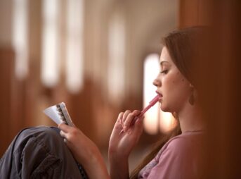 A young woman ponders ideas while holding a pen and notebook indoors, evoking contemplation.