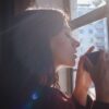 A woman savors a hot drink by a sunlit window in an urban setting.
