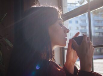 A woman savors a hot drink by a sunlit window in an urban setting.