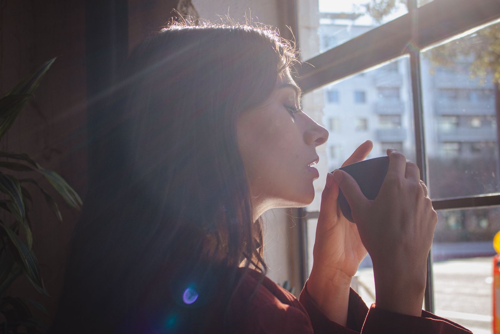 A woman savors a hot drink by a sunlit window in an urban setting.