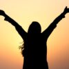 Silhouette of a woman with raised arms against a vibrant sunset sky, symbolizing freedom and joy.