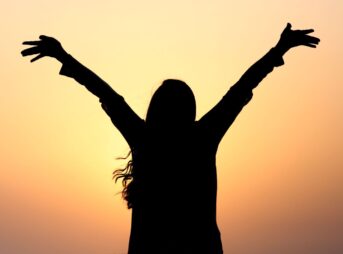 Silhouette of a woman with raised arms against a vibrant sunset sky, symbolizing freedom and joy.