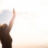 A woman with raised arms enjoying the bright open sky in a serene outdoor setting.