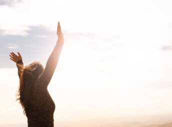 A woman with raised arms enjoying the bright open sky in a serene outdoor setting.