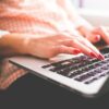 Close-up of a woman with red nails typing on a laptop indoors.