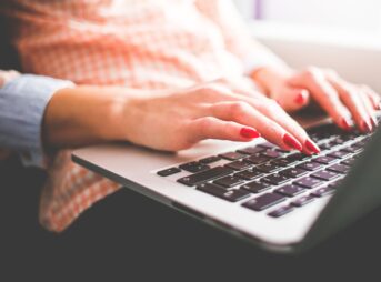 Close-up of a woman with red nails typing on a laptop indoors.