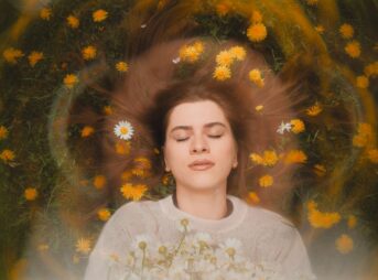 Relaxed woman with closed eyes, surrounded by daisies and dandelions, embracing nature's beauty.