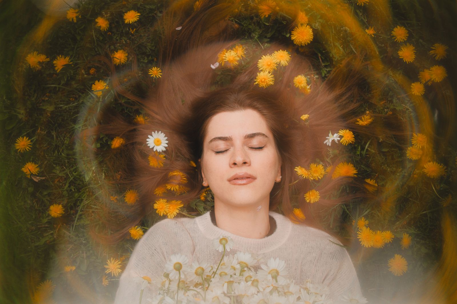Relaxed woman with closed eyes, surrounded by daisies and dandelions, embracing nature's beauty.