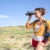 Young woman enjoying a sunny day hike, using binoculars to explore nature and capture the scenic view.