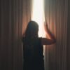 Back view of anonymous lady opening ribbed curtain behind window with glowing light while standing in apartment