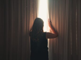 Back view of anonymous lady opening ribbed curtain behind window with glowing light while standing in apartment
