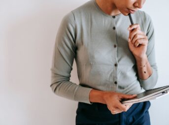 Crop unrecognizable female worker in casual clothes standing with clipboard and notebook and touching chin with pen against white wall