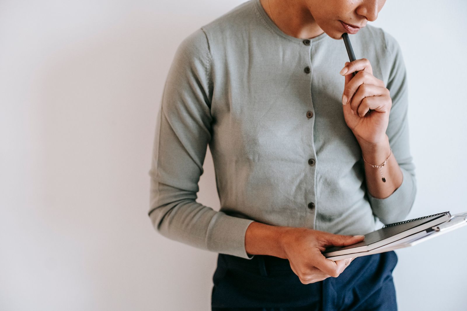 Crop unrecognizable female worker in casual clothes standing with clipboard and notebook and touching chin with pen against white wall