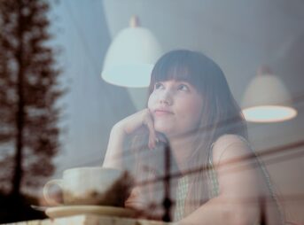 Thoughtful woman gazing outside a café window with reflections and warm lighting.