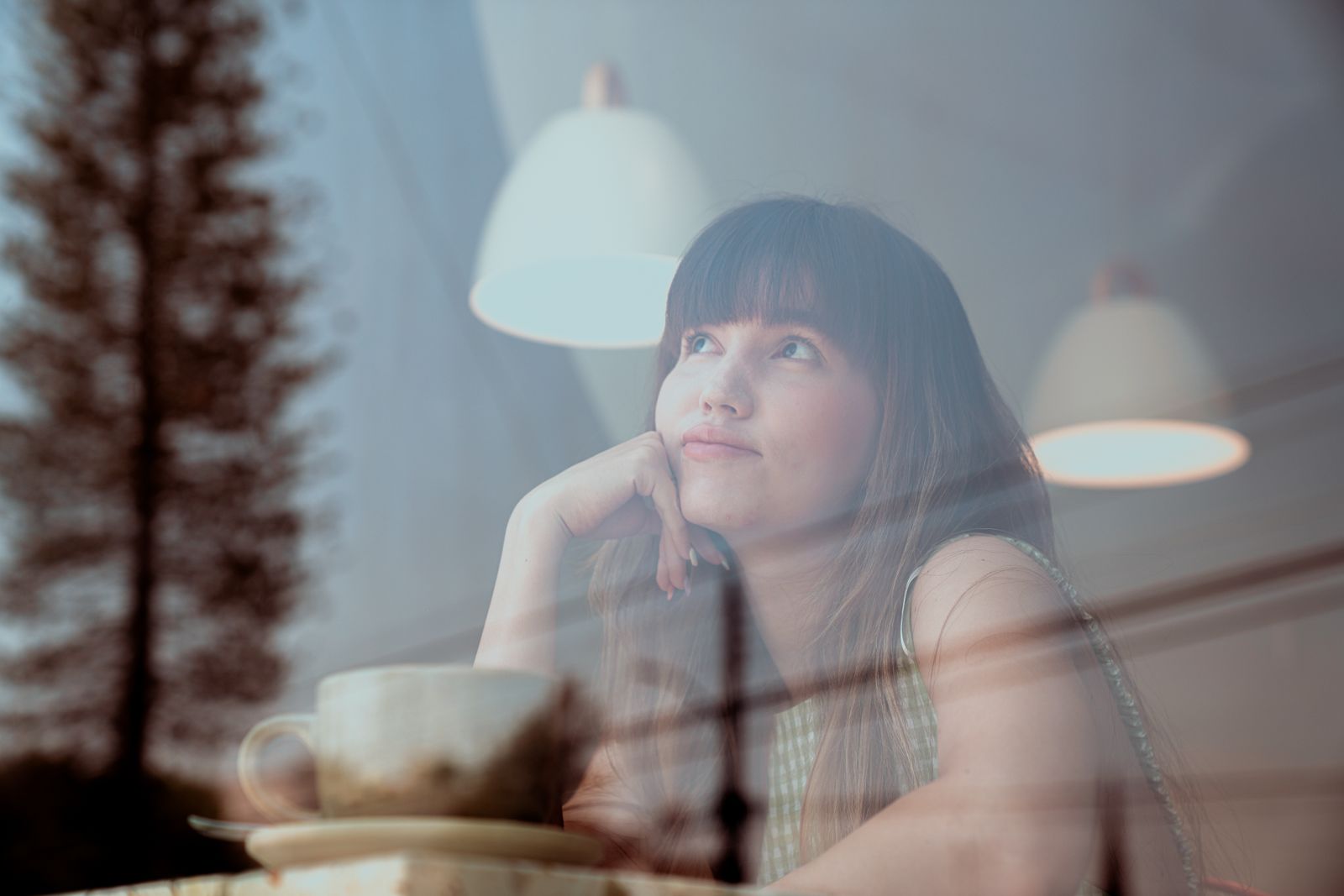 Thoughtful woman gazing outside a café window with reflections and warm lighting.