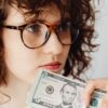 Close-up of a woman holding a US five dollar bill, wearing glasses and looking thoughtful.