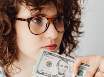Close-up of a woman holding a US five dollar bill, wearing glasses and looking thoughtful.
