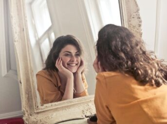 A cheerful woman smiles at her reflection in a vintage-style mirror, exuding positivity and warmth.