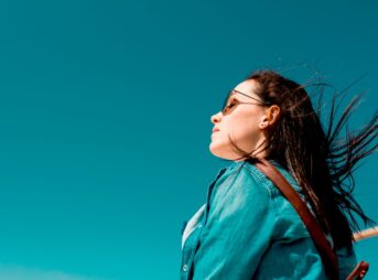 Fashionable woman outdoors in denim shirt with sunglasses under clear blue sky, enjoying sunny weather.