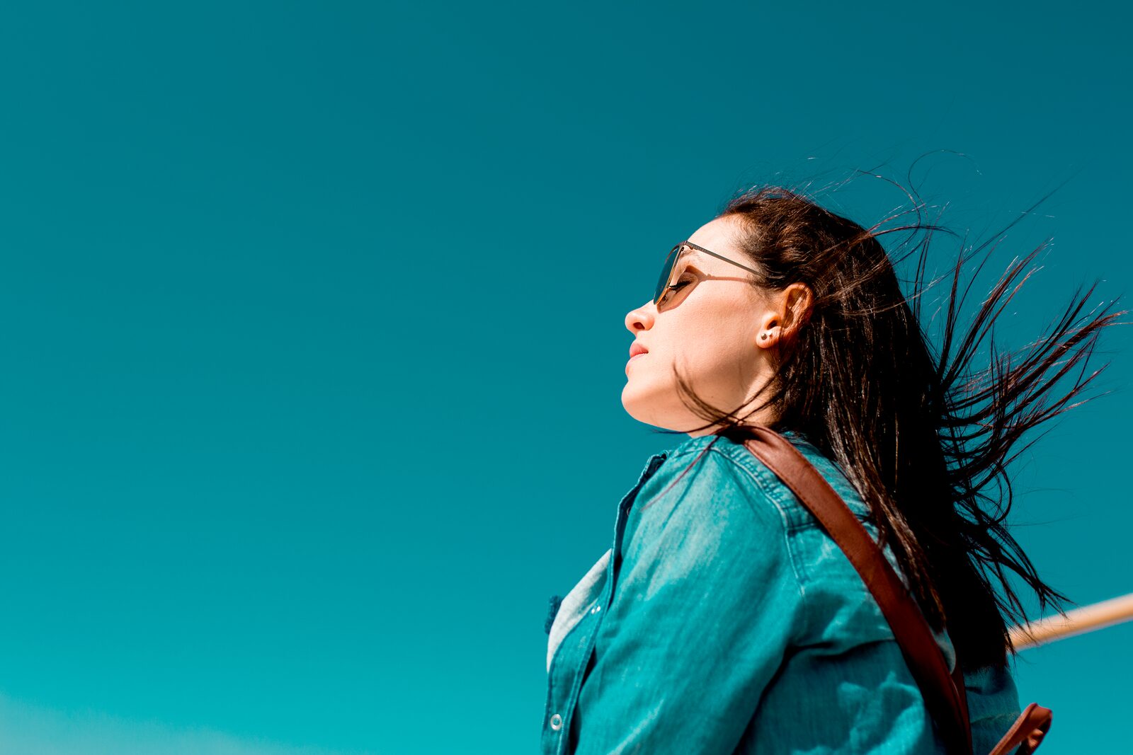Fashionable woman outdoors in denim shirt with sunglasses under clear blue sky, enjoying sunny weather.