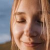 Close-up of a smiling woman with closed eyes, sunlit hair, and a serene expression outdoors.