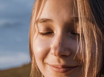 Close-up of a smiling woman with closed eyes, sunlit hair, and a serene expression outdoors.