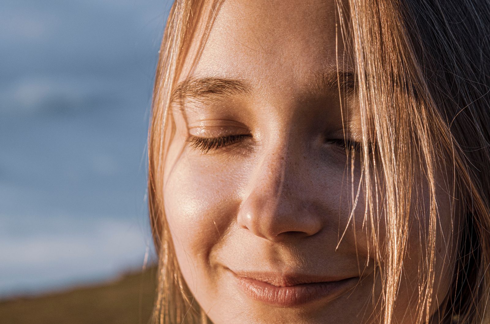 Close-up of a smiling woman with closed eyes, sunlit hair, and a serene expression outdoors.