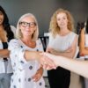 Diverse group of women in an office setting shaking hands and smiling, symbolizing partnership.