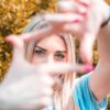 A young woman frames her face with hands, focusing on her eyes, in a vibrant outdoor setting.