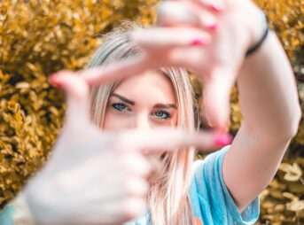 A young woman frames her face with hands, focusing on her eyes, in a vibrant outdoor setting.