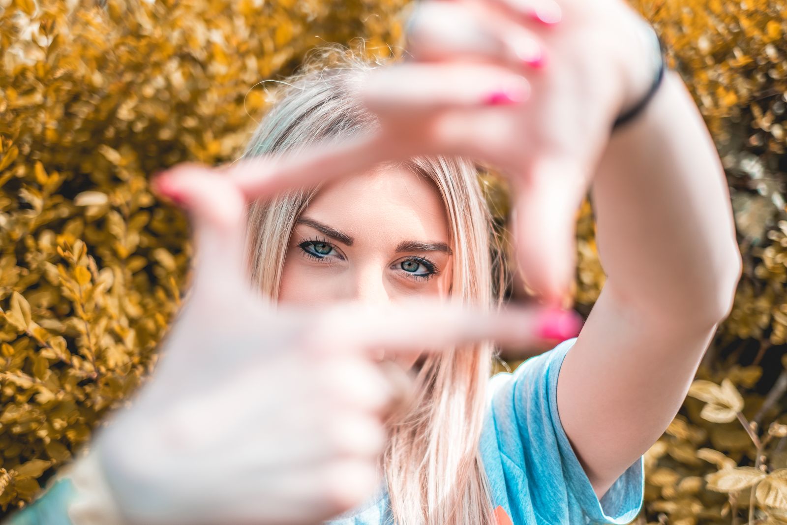 A young woman frames her face with hands, focusing on her eyes, in a vibrant outdoor setting.