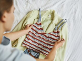 Woman arranging a stylish blazer and knitted top outfit on a bed.