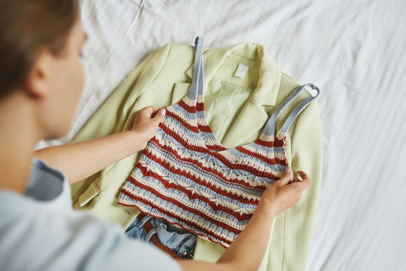 Woman arranging a stylish blazer and knitted top outfit on a bed.