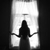 grayscale photo of woman in dress standing near window
