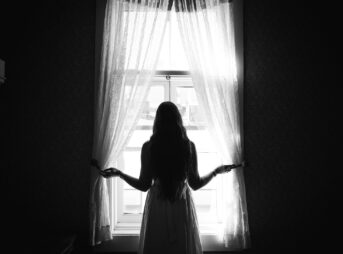 grayscale photo of woman in dress standing near window